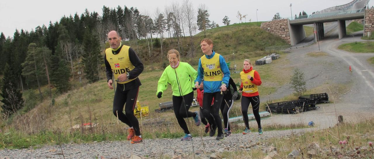 En gruppe på fem løper oppover bakken. Foran løper en ledsager med gul refleksvest merket «guide»