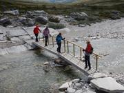 Noen i turfølget krysser en smal bro over en fjellelv